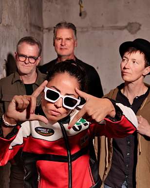 La band ask Robin davanti a un muro di cemento: la frontwoman Regina Brury, con giacca da moto rossa e occhiali da sole bianchi, forma una cornice con le mani, dietro di lei tre membri della band.