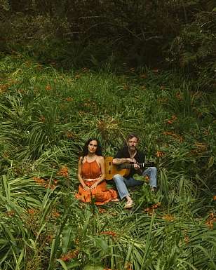 La chanteuse Lea Lu, en robe orange, et un guitariste avec une guitare classique sont assis dans une végétation haute et dense, parmi des fleurs orange.