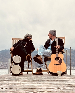 Zwei Musiker von Mar Y Sol sitzen auf einem Holzsteg am See, einer mit Gitarre, der andere mit Bongos, und unterhalten sich entspannt vor einer Bergkulisse.