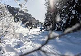 Deux fondeurs tracent leurs traces sur la piste enneigée en Engadine, avec un hôtel de montagne historique en arrière-plan parmi les sapins enneigés.