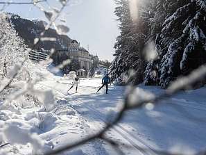 Cross-country skiers gliding along the winter trail in the Engadin, surrounded by snow-covered fir trees and a historic hotel backdrop