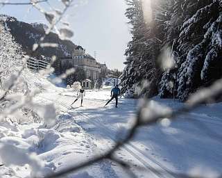 Two cross-country skiers on a trail in the Engadin, with a historic hotel building in the background, framed by snow-covered branches and fir trees