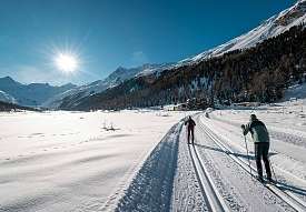 Due sciatori di fondo su una larga pista in Engadina con vista panoramica sulla valle e sole invernale brillante.