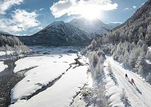 Zwei Langläufer auf einer Loipe entlang eines verschneiten Flussufers in Pontresina mit Bergpanorama und Sonnenstrahlen.