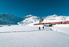 Due escursionisti con le ciaspole avanzano nella neve vicino a Pontresina, mentre il Trenino rosso del Bernina passa davanti alle cime innevate.