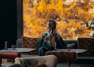 Frau mit einer Tasse in der Hand sitzt in einer Hotellounge mit Sesseln und Holztischen und blickt durch ein grosses Fenster auf goldgelbe Lärchen