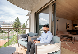 Homme travaillant sur un ordinateur portable sur le balcon d’une chambre du Sunstar Hotel Pontresina avec vue sur les montagnes et le paysage de l’Engadine.