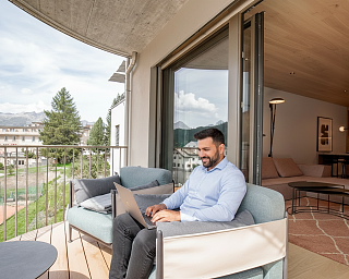 Homme travaillant sur un ordinateur portable sur le balcon d’une chambre du Sunstar Hotel Pontresina avec vue sur les montagnes et le paysage de l’Engadine.